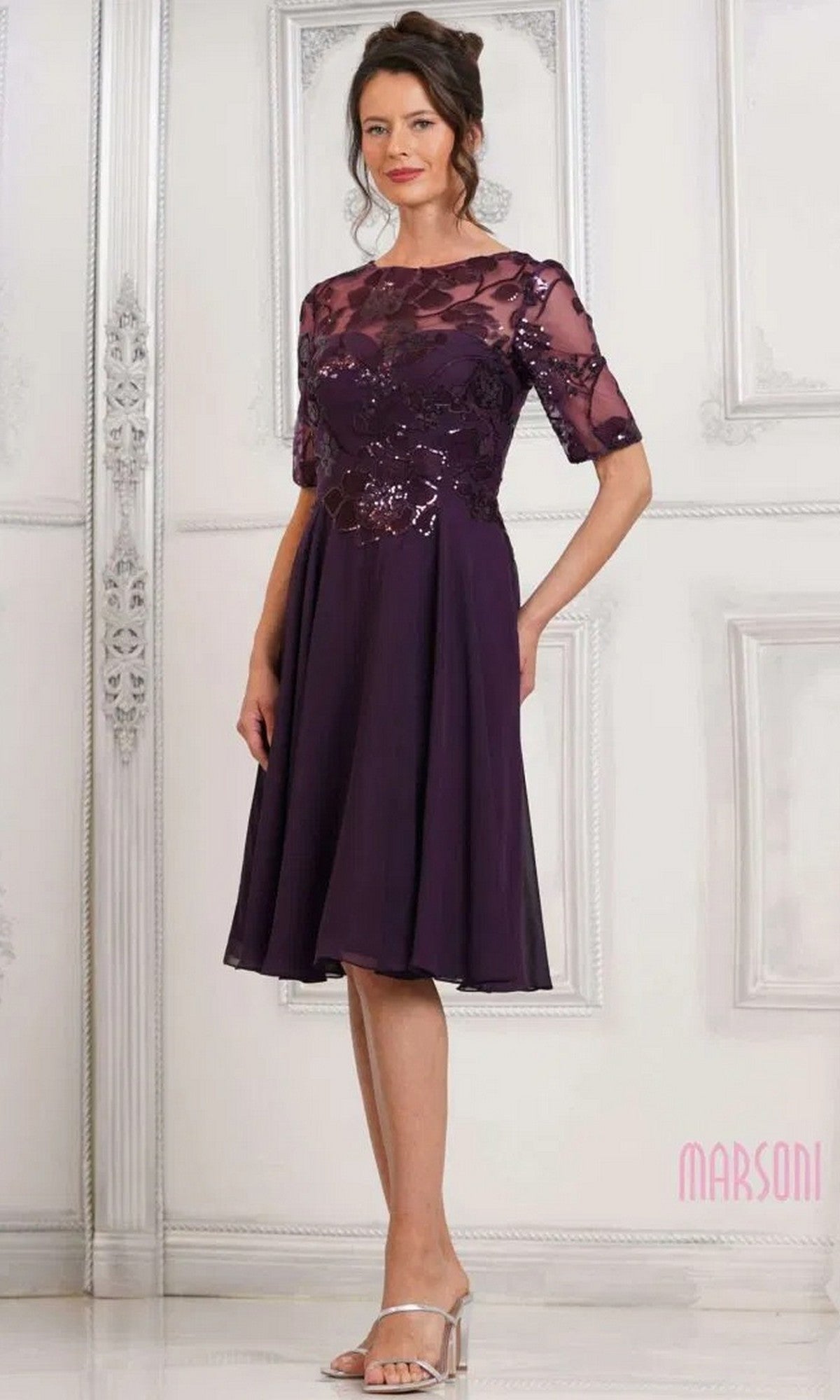 A woman stands in a light room with ornate wall panels, wearing the Marsoni M286S semi-formal knee-length dress in eggplant, featuring floral sequin lace sleeves and bodice with an elegant A-line chiffon skirt.